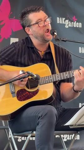 A man with short dark hair, glasses, and a goatee is singing and playing an acoustic guitar. He is wearing a black and gray striped shirt. He is seated on a folding chair. In the background, there is a banner with a large pink butterfly and the words "BAKgården" repeated. Microphones are positioned in front of the singer and his guitar. A music stand is visible to the right of the singer.