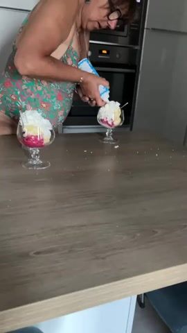 A middle-aged woman with glasses and dark hair, wearing a floral-patterned slip dress, is in a kitchen. She is standing at a wooden table and is in the process of adding whipped cream from a can onto two dessert dishes. The dishes contain layers of what appears to be cake, possibly fruit, and whipped cream. The woman is focused on the dessert in front of her, squeezing the can of whipped cream onto the top. In the background, a microwave and kitchen cabinets are visible. The lighting is bright, suggesting daytime.