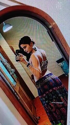 A woman is taking a selfie in a mirror. She is wearing a patterned top and skirt. The background appears to be an indoor setting.