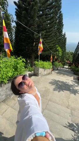 A person with sunglasses and a white hoodie walks up a stone staircase lined with trees and flags. The person is filmed from a first-person perspective, and the camera pans up to show the staircase and the surrounding environment. The staircase appears to be part of a temple or religious site, as suggested by the flags and the architectural style of the stairs and railings. The weather is sunny, and the sky is clear blue. The video captures the upward movement on the stairs and the beautiful scenery of the location.