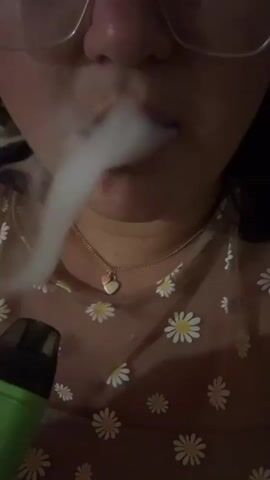 This is a close-up, low-angle shot of a person's face as they exhale a cloud of vapor from their mouth. The person is wearing glasses and a necklace with a heart-shaped pendant. Their shirt has a daisy pattern. In the lower-left corner of the frame, a green vaping device is visible. The lighting is dim.