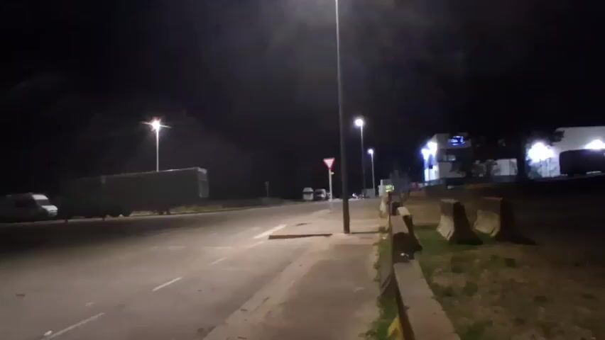 This is a nighttime video taken from a road. The scene is dimly lit by streetlights. There is a paved road with white markings and a sidewalk on the right. On the left side of the road, there are some trees and bushes. In the distance, there are some buildings and more trees.

The main subjects in the video are several vehicles and a group of people. On the left side of the road, a large, green trailer is parked, and a white van is visible behind it. Further down the road, a white van and another small vehicle are parked. In the center of the frame, near a yield sign, there is a bright red light, possibly from a flare or a vehicle's brake lights, and a group of people are gathered around it. One person appears to be holding a bright red object that is emitting smoke or sparks. The people are mostly silhouetted against the light, making it difficult to discern their features or clothing. They seem to be interacting with each other or observing the bright red light.

The overall atmosphere is somewhat mysterious due to the low light conditions and the presence of the unexplained red light and smoke. The time of day is clearly night, indicated by the dark sky and the artificial lighting. There are no obvious weather cues, but the scene appears calm. The presence of streetlights, a yield sign, and concrete barriers suggests an urban or semi-urban environment, possibly near a road junction or an industrial area. The location context provided is Mas Aliu, Spain. No visible text is present in the video.
