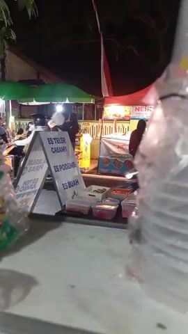 The video shows a night market scene. In the foreground, there are plastic-wrapped items, possibly food or merchandise. In the middle ground, an A-frame sign displays menu items in Indonesian, including "ES TELER CREAMY", "ES PODENG", and "ES BUAH". There are containers of ingredients behind the sign. Several people are visible in the background, some near food stalls with green and red awnings, and others on motorcycles. One person is seated on a motorcycle. An Indonesian flag is partially visible hanging from a pole. The overall atmosphere appears to be a lively street food vendor area.