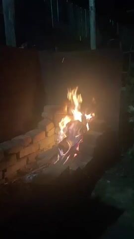 The video begins in darkness, with a fire burning in a makeshift brick structure. The flames are bright orange and yellow, with hints of blue and purple at the base, indicating intense heat. The brick structure appears to be a rudimentary fireplace or fire pit, with several layers of bricks forming an enclosure for the fire. The background is dark and out of focus, suggesting it is nighttime. As the video progresses, the camera pans slightly to the left, revealing more of the dark environment. There are faint outlines of what appear to be wooden structures or fencing in the background. The fire continues to burn, casting a warm glow on the surrounding bricks and creating dancing shadows. The scene is quiet, with no discernible sounds other than the crackling of the fire. The overall impression is one of a calm, solitary moment outdoors, likely in a backyard or a secluded area.