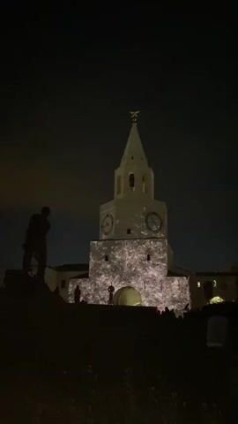 At night, a white tower with a steeple and two clock faces is illuminated with a light show. Silhouetted figures of people stand below the tower, likely watching the light show. To the left, a dark statue is visible against the night sky. The overall scene is dark, with the tower and its projections being the main source of light.