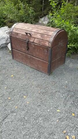 A handheld camera pans from left to right, focusing on a wooden chest. The chest is brown with metal banding and a latch. It is resting on a gravel surface, with foliage and rocks visible in the background. A person's hand enters the frame and begins to manipulate the latch on the chest. The video ends with the latch still being operated.