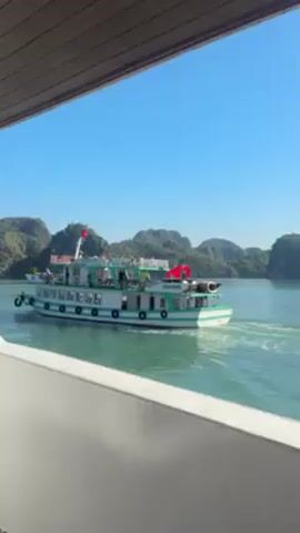 The video shows a white and green tourist boat moving through water. The boat has several red flags on it. In the background, there are islands covered in vegetation. The sky is blue. The video is taken from another boat, looking out over the water. A person is heard talking in the background.
