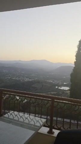 The video pans across a scenic landscape viewed from a balcony. In the foreground, a dark-colored chair is partially visible. A balcony railing with an ornate design runs horizontally across the frame. Beyond the balcony, a vast expanse of rolling hills covered in greenery stretches out towards a body of water. Mountains are visible in the distance, softened by a hazy atmosphere. A tall, slender cypress tree stands prominently to the right of the frame, its silhouette stark against the pale, possibly dusk or dawn, sky. The overall scene depicts a tranquil, natural environment.