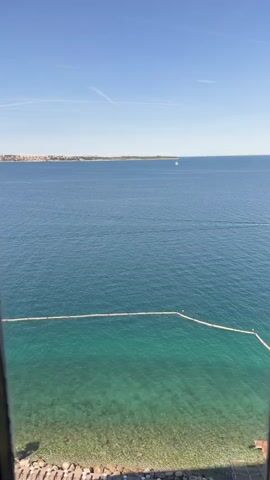 A video begins by showing a clear blue sky and the calm surface of the sea. The camera pans down to reveal a section of clear turquoise water with a rocky bottom visible. A long, white floatation device runs parallel to the shoreline, separating the shallow, clear water from the deeper, darker blue ocean. The shoreline itself is made up of rocks and some visible vegetation. The video then abruptly cuts to a dark, out-of-focus image, followed by a blurry shot of a light-colored wall. The video appears to be filmed from an elevated position, possibly a window or balcony.