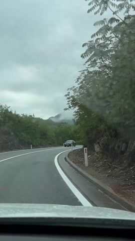 The video captures a first-person perspective from inside a vehicle driving on a winding, two-lane asphalt road. The road is marked with white lines, and appears wet from rain or recent precipitation. Lush green trees and vegetation line both sides of the road, forming dense foliage, particularly on the right. In the background, mountains are visible, partially obscured by low-hanging clouds and fog under an overcast sky.

Raindrops are visible on the vehicle's windshield throughout the recording. Several vehicles are seen passing in the opposite direction: a dark-colored sedan at the beginning, followed by a red MAN flatbed truck carrying large, dark rectangular objects (possibly metal beams), and later a yellow/gold car.

Notable road signs include a white rectangular sign displaying "M-23 km 14" (indicating Route M-23, kilometer 14) and, further along, a circular warning sign for a steep downgrade, positioned above a circular speed limit sign indicating "80" km/h. White marker posts line the right edge of the road.

The scene depicts a scenic drive through a mountainous and verdant region, consistent with the location of Gospoština, Montenegro, during what appears to be an overcast or rainy day.