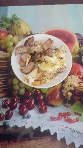 A plate of food sits in the center of a table with a colorful fruit-themed placemat. The plate contains pieces of cooked meat and scrambled eggs. Surrounding the plate on the placemat are depictions of grapes, cherries, red currants, pears, watermelon, and other fruits. The overall scene suggests a meal, possibly dinner.