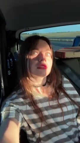 A young woman with long brown hair and red lipstick sits in the passenger seat of a vehicle. Sunlight streams through the window, casting stripes of light and shadow across her face and striped shirt. She turns her head to look around, her eyes darting from side to side before she looks directly at the camera. The background shows a blurred landscape outside the car window.