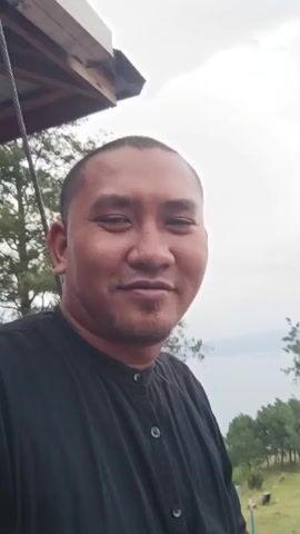 The video opens with a close-up shot of a man with a shaved head and a goatee, wearing a black collared shirt. He is smiling at the camera. The background is slightly out of focus, but it appears to be an outdoor setting with a body of water in the distance, possibly a lake, and lush green hills. As the video progresses, the camera pans slightly to the right, revealing more of the environment. There are trees, some with bare branches, and a clear sky with scattered clouds. Further in the background, a transparent walkway or bridge is visible, with several people walking on it. Some of these individuals are wearing headscarves. There are also some decorative elements in the landscape, including what appears to be a crescent moon sculpture and some colorful flowers. The man in the foreground remains the primary subject, but the background shows a scenic view and other people enjoying the location. The overall impression is of a tourist spot with a beautiful natural backdrop and an elevated walkway offering panoramic views.