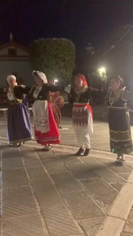 A group of five people are performing a dance outdoors at night. Four of the dancers are women dressed in traditional Greek folk costumes. The first woman from the left wears a dark jacket, a white headdress, a blue skirt, and black shoes. The second woman wears a dark jacket, a white headdress, a white apron over a red skirt, and white shoes. The third woman wears a dark jacket, a red headscarf, a white apron over loose white pants, and dark shoes. The fourth woman wears a dark jacket, a dark skirt with gold trim, and dark shoes. The fifth person, on the far right, is partially visible, also wearing a dark garment. The dancers are arranged in a line, facing towards the viewer and extending their arms. The background shows a building with a red roof and white walls, lush green bushes, and streetlights illuminating the stone-paved area where the dance is taking place. The ground is paved with large, rectangular stones.