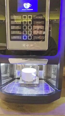 The video shows a Necta coffee machine dispensing a mocaccino into a white ceramic mug. The machine is black with blue lights illuminating the dispensing area. The control panel shows various coffee options in French. The background reflection suggests someone is operating the machine. The mug is placed on a metal grate within the machine. The surrounding counter is wooden.
