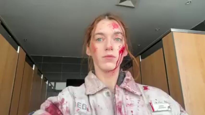 A person with reddish-brown hair and light eyes stands in what appears to be a restroom. They are wearing a light-colored jacket with a name tag reading "Emma Johnson". Their face and jacket are covered in what appears to be fake blood. The restroom has light wood-paneled stalls and a dark grey ceiling. The lighting is bright and somewhat clinical. The person looks directly at the camera with a neutral expression.