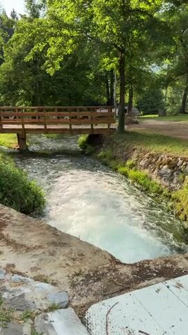 This is a vertical video of a stream flowing under a wooden footbridge in a park-like setting. The water is churning and white, suggesting it is moving quickly. Lush green trees and foliage surround the stream, with sunlight dappling through the leaves. The foreground shows a rough stone embankment and a wooden plank. The scene appears tranquil and natural.