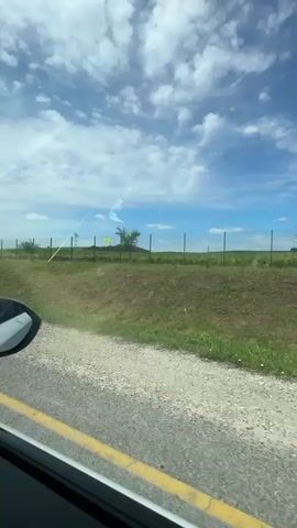 The video shows a view from a car window looking out at a rural landscape. The car is on a paved road with a yellow line on the left side. To the right of the road, there is a gravel shoulder, then a grassy area with some small trees and a wire fence. Beyond the fence, there is a field of grapevines and a gentle hill in the distance. The sky is blue with white clouds, and there appear to be contrails from an airplane visible. The video is short and appears to be a travel or scenery clip.