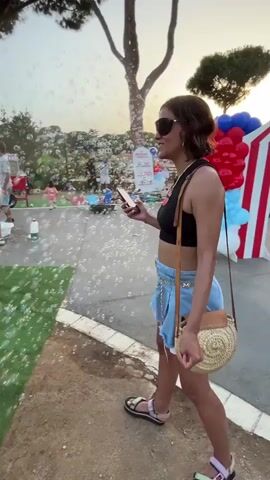 A woman in sunglasses and a black crop top with a blue denim skirt stands on a dirt path and records a video on her phone. She is wearing platform sandals and carrying a straw purse. Bubbles are floating in the air around her. In the background, other people are gathered on a green lawn, some of whom appear to be watching or participating in bubble-blowing activities. Trees and a red, white, and blue balloon arch are also visible in the background. The scene appears to be outdoors, possibly at a park or event.
