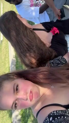 The video shows a group of people outdoors. The camera is oriented vertically and appears to be handheld. In the foreground, a young woman with long brown hair is looking into the camera. She has bright pink blush on her cheeks and is wearing makeup. Her hair is partially obscuring her face. Behind her, another young woman with long brown hair is visible. She is wearing a black top with a floral pattern. Further back, a man wearing a black baseball cap and a white t-shirt is smiling and looking towards the camera. Another person is partially visible in the background. The setting appears to be a grassy area with a paved pathway or walkway. The lighting suggests it is daytime, possibly sunny.