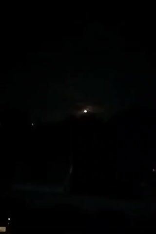 The video depicts a nighttime scene. The moon is visible through the clouds in the sky, but it is obscured. Below the sky, the silhouettes of trees and rooftops are visible. In the foreground, a car with its parking lights on is parked on what seems to be a street. The scene is very dark.
