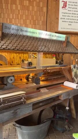 The video shows a close-up, steady shot of a model of an old saw, labeled "Alte Säge Heimenhausen 1807 / Modell im Massstab 1:10." The model is intricate and appears to be made of wood and metal. It is housed in a structure with a shingled roof, reminiscent of a traditional building. Below the model, a workbench is visible with various items, including a roll of paper, a stack of wood, and a metal tub. A vacuum cleaner is also visible on the right side of the workbench. In the background, a wooden wall is covered with square tiles, and a framed text in German is mounted on it. The text appears to be a biblical quote. The lighting is warm and illuminates the model and its surroundings. The overall impression is of a detailed and well-maintained display of a historical piece of machinery.