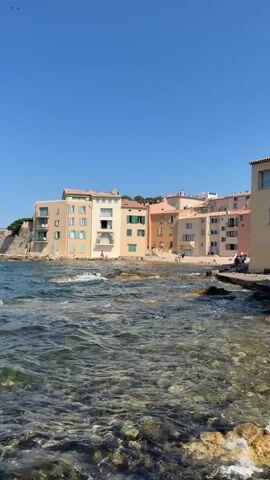 The video shows a waterfront view of a town with pastel-colored buildings. The water in the foreground is clear, revealing rocks and seaweed beneath the surface. Waves gently lap at a rocky shoreline. In the background, several people can be seen walking along a path in front of the buildings. The sky is clear and blue.