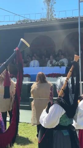 This video captures a ceremonial event outdoors on a sunny day, likely a historical reenactment or festival. In the foreground, two individuals are partially visible, facing away from the camera, playing bagpipes. The bagpipes on the left are black with a red and black tasseled bag, and the bagpipes on the right, played by a person in traditional attire including a patterned scarf and a dark skirt, are also black with visible wooden pipes and a bag.

Behind the bagpipers, a stage area is set up. Several people are seated on the stage behind a long table draped with a blue cloth and decorated with red flowers. Some individuals on the stage appear to be wearing white or light-colored robes, suggesting a formal or religious aspect to the event. A banner with a circular, red, patterned design is visible on the backdrop behind the seated individuals. The setting appears to be grassy, with some sort of structure or platform forming the stage. Sunlight is bright, casting shadows and creating lens flare effects in the upper portion of the video. The overall atmosphere suggests a communal gathering with a focus on traditional music and ceremony.