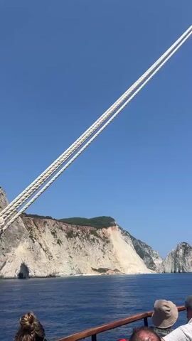 The video shows a view from the deck of a boat looking out towards a coastline with white cliffs. There are several people on the deck, some of whom are looking at the cliffs. One person is taking a photo of the cliffs with their phone. The sky is clear and blue, and the sea is calm. The boat appears to be sailing.
