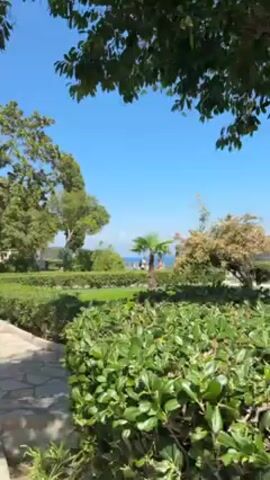 The camera pans across a garden on a clear, sunny day. There are various trees, manicured hedges, and a stone pathway. In one area, several people sit on a bench under a large tree. In the background, one can see the ocean.