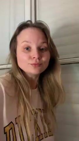 A woman with blonde hair wearing a white sweatshirt with a yellow letter on it stands in front of a window with blinds. She smiles, then turns to face a tricolor cat. The woman speaks to the cat, then puts her hand to her ear like a telephone. She gestures her hand to her ear again, as if talking on the phone.