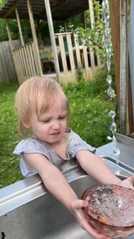 A young child with blonde hair, wearing a dirty grey t-shirt, stands at a metal sink outdoors. The child is holding a clear glass bowl in both hands under a stream of running water. The child’s facial expression is one of mild displeasure or discomfort. Water is splashing around the bowl and on the child’s hands and arms. In the background, there is lush green grass, a wooden structure with a roof, and some trees.