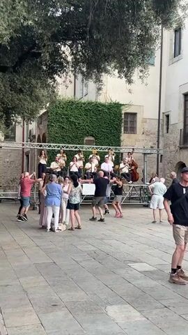 A band plays music on a raised stage in an outdoor courtyard. Several people are dancing in front of the stage, and other people are watching. The courtyard is paved with cobblestones and surrounded by buildings, one of which is covered in ivy. Trees provide shade over a portion of the courtyard. The band members are dressed in light-colored outfits and some are playing brass instruments. The dancers are dressed in casual clothing. The overall atmosphere appears festive.