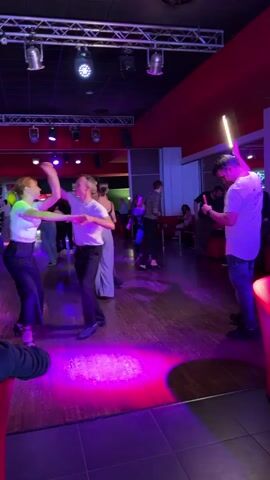 The video captures an energetic indoor scene at what appears to be a dance club or studio in Buccinasco, Italy. Multiple people are engaged in social dancing on a wooden floor under dim, colorful lighting, primarily purple and blue, emanating from spotlights on a metal truss structure above. The setting features a red wall on the right and dark ceilings.

In the foreground, a couple dances in synchronized steps, while another woman dances nearby. To their right, a man in a white T-shirt and jeans holds a glowing object, possibly a phone or light stick, and appears to be recording or interacting with it, briefly giving a thumbs-up gesture. In the background, other couples and individuals are also dancing or mingling. The atmosphere suggests a lively dance event, possibly a salsa or bachata social, with participants dressed in casual attire. The overall mood is active and joyful, indicative of a social gathering focused on dance. No specific text is visible in the frame.