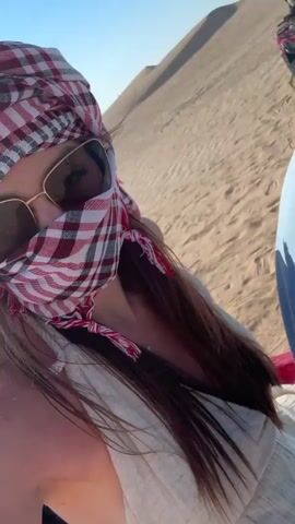 The video shows a group of people getting ready to ride ATVs in a desert environment. The camera appears to be held by a person wearing sunglasses and a red and white checkered headscarf, sitting on a red ATV.  Another person, wearing a blue shirt and black and white checkered headscarf, is sitting next to them. In the background, other ATVs are visible, some with people already riding them up a sand dune under a clear, sunny sky. The sand is textured and dry. The ATVs are red.