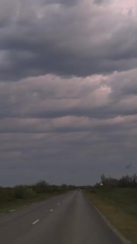 The video shows a view of a road leading into the distance under a cloudy sky. The first shot is a straight view down a rural highway with sparse vegetation on either side. The sky is predominantly grey clouds. The video transitions to a view from inside a moving vehicle. The camera pans up to show the sky as seen through a window. The sky is now a mix of grey, white, and pink clouds, with a thin strip of yellow indicating the setting sun on the horizon. The landscape is blurred due to the motion of the vehicle.