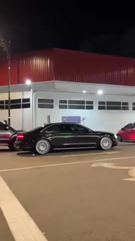At night, a dark Audi car sits still at the side of a road next to a tall building with a white facade and a red roof. A tall, ornate lamppost stands on the road near the front of the car. Other cars, including a silver one, move on the road.