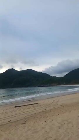 A drone shot pans across a tropical beach at dusk. The sky is cloudy and overcast, with the sun setting behind a large, dark green, jungle-covered mountain range that dominates the background. The ocean is a vibrant turquoise, with gentle waves breaking on the shore. A few people can be seen swimming and wading in the water. The beach itself is made of light brown sand, with tire tracks and a piece of driftwood visible. The overall impression is one of a peaceful and serene evening at a secluded tropical paradise.