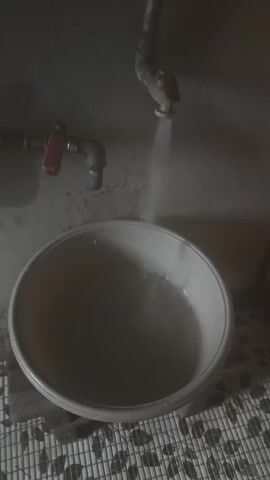 A faucet is dripping water into a basin. The basin is full of water. The faucet is located on the wall. The wall is gray. The floor is tiled.