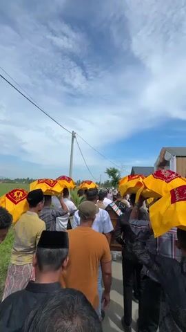 The video shows a procession of men walking along a dirt road under a partly cloudy sky. They are carrying what appear to be covered objects above their heads, held by umbrellas or canopies. The canopies are yellow with red trim and intricate designs. Many of the men are wearing traditional attire, including sarongs and peci hats. The environment appears rural, with a field on the left side of the road and some buildings on the right. The perspective is from behind the procession, looking forward. The men are walking away from the camera.