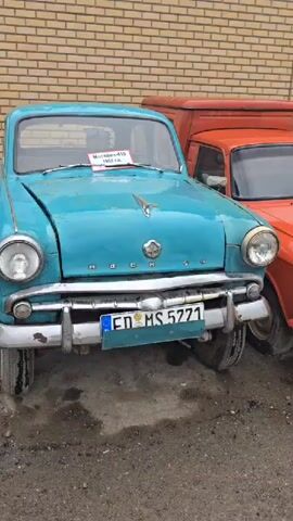 The video pans across a row of vintage cars parked next to a brick building. The cars are various colors, including turquoise, orange, yellow, blue, red, and green. Some of the cars have license plates visible. The ground is wet and appears to be asphalt or gravel. The lighting suggests it is daytime, possibly overcast. There are no people visible in the video. The overall impression is of a car show or a collection of classic vehicles.