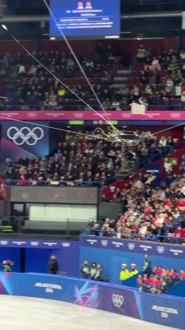 A couple is skating on a rink that is decorated with the Olympic rings and Milano Cortina 2026 branding, with a crowd in the background. A camera crane is visible above the rink. The woman skater wears a dress with a brown and gold pattern and her partner is in a black suit. At the end of the video, both skaters raise their hands in the air.