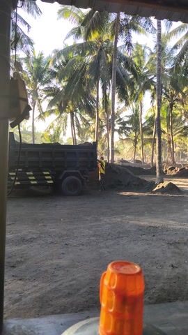 The video shows a scene in Ijobalit, Indonesia, featuring a truck, palm trees, and a person. The truck appears to be a dump truck, dark in color with yellow markings along its side. A person wearing a yellow vest is visible near the truck, possibly involved in loading or unloading it. The background is dominated by a dense cluster of tall palm trees, their fronds rustling gently in what seems to be a warm, sunny climate. The lighting suggests it is either early morning or late afternoon, with the sun casting long shadows. The ground is rough and dusty, indicative of a rural or industrial area. An out-of-focus orange object, likely a bottle or container, is prominently placed in the foreground, partially obscuring the view of the scene beyond. The overall impression is of a working day in a tropical environment.