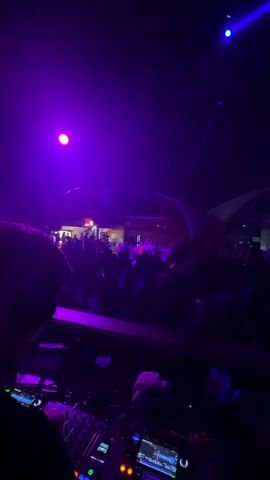 The video shows a crowded nightclub from the DJ’s perspective. A DJ console is in the foreground, with a crowd of people dancing in the background. The club is lit with bright purple and blue lights. Some balloons and architectural elements can be seen in the background. The camera appears to be moving slightly.