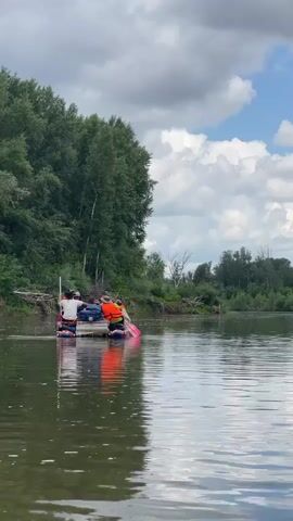 Four people are on a raft on a river. Three of the people are wearing life vests, one orange, and two blue. Two of the people are holding paddles. One person is holding a long pole. The raft is made of logs. The river is surrounded by trees on the left and right. The sky is cloudy.