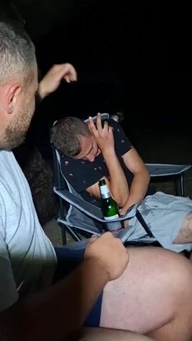 Two men are sitting outside in the dark. One man, wearing a grey t-shirt, is in the foreground, facing away from the camera. The other man, wearing a black t-shirt with a red pattern, is seated in a camping chair in the background, leaning his head down and touching his forehead. He appears to be looking at a green glass bottle with a white label that is placed in a holder on the side of his chair. The man in the foreground has his left hand resting on his lap and his right hand is not visible. The man in the background has his right arm bent and his hand near his face, and his left arm is resting on the armrest of the chair. The lighting is dim, suggesting it is nighttime. The environment appears to be outdoors, possibly a campsite.