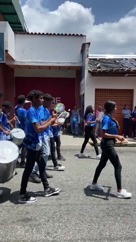 A group of approximately a dozen young men and women are parading down a paved street, playing drums and tambourines. They are wearing matching bright blue t-shirts with a white logo and graphic designs, with most wearing jeans or dark pants and sneakers. The street is lined with buildings of brick and whitewashed cement, with painted store fronts and shuttered windows. The sky is cloudy with a few patches of bright blue visible.