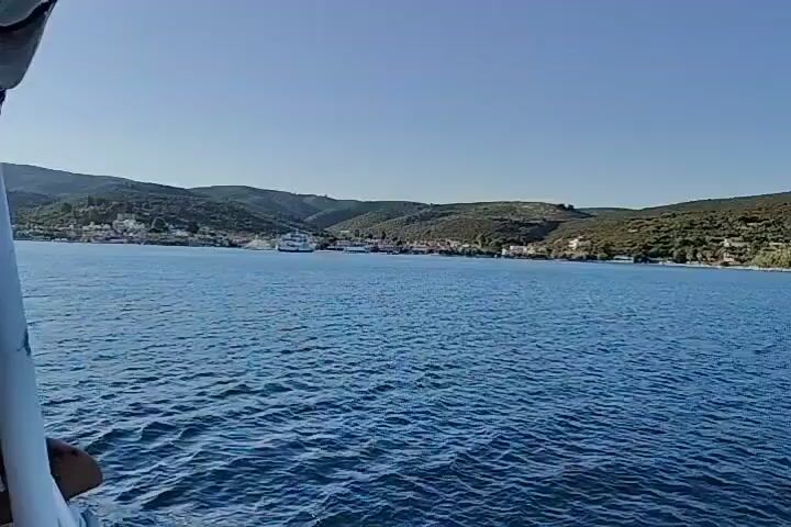 The video captures a view from a moving boat on a clear, sunny day. The water is a vibrant blue, with gentle ripples indicating calm conditions. In the distance, a coastal town is visible, nestled at the base of rolling, green hills. The town features a line of buildings, some with red-tiled roofs, and a prominent white ferry docked in the harbor. The ferry appears to be a significant structure, suggesting a transport hub. The hills are covered in dense vegetation, with some patches of lighter green suggesting scrub or dry grass. The sky is a clear, bright blue, with no clouds visible. The camera perspective is from a low angle, looking out over the water towards the shore. The edge of the boat, likely a railing or part of the vessel, is visible in the foreground on the left side of the frame, partially obscuring the view. The movement of the boat creates a slight parallax effect as the landscape recedes.