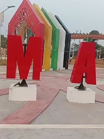 The video displays a large, red, three-dimensional letter 'M' followed by a large, red, three-dimensional letter 'A', both mounted on white blocks and set against a background of colorful pillars. The scene appears to be an outdoor public space, possibly a park or plaza, with paved ground featuring red and grey patterns. In the distance, a wooden structure with a curved roof is visible, along with trees and other decorative elements. The lighting suggests it is daytime, possibly overcast. The text "Taman Kota" (City Park) and "Keren Bangkrai" (Cool Bangkrai) appear overlaid on the video, indicating the location or a description of the place. The camera is panning across the letters, revealing more of the environment.