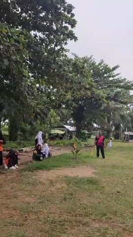 A group of people are gathered in an outdoor grassy area. A man in a red shirt and black pants stands facing left, holding a yellow stick. To his left, a young person in a green shirt and black pants is throwing something. Further left, several other people, including some wearing headscarves, are sitting on the ground. Trees are visible in the background, along with some structures that appear to be small buildings or stalls. The sky is overcast.